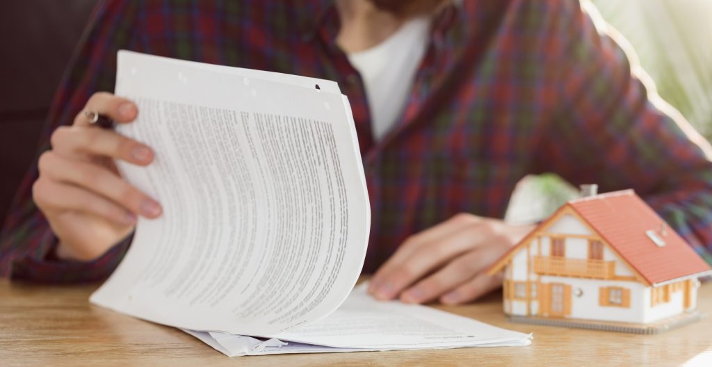 A person in a plaid shirt reviewing paper documents on a wooden desk next to a small model house.