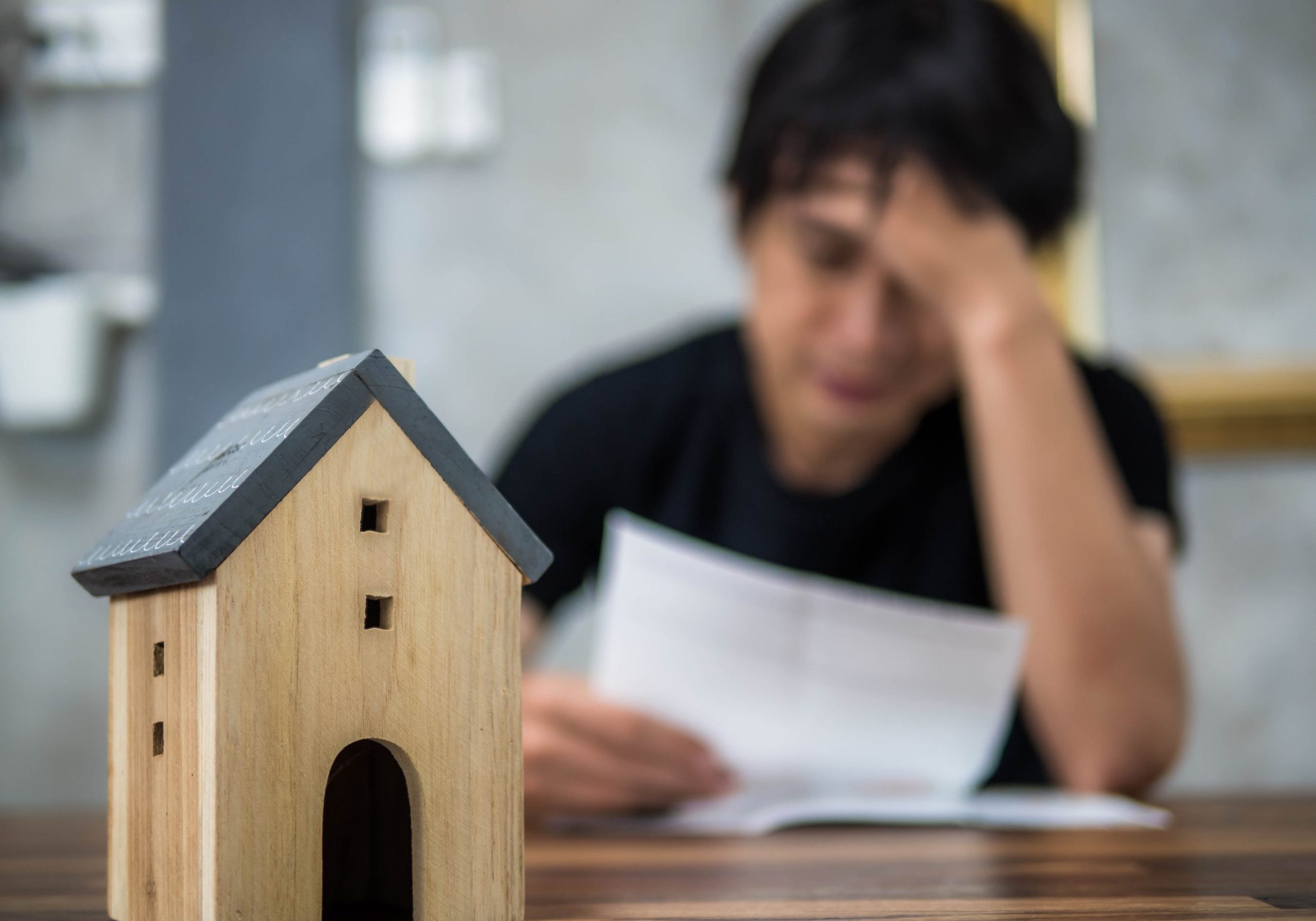 Stressed man sitting at a desk with a house model, overwhelmed by piles of bills.